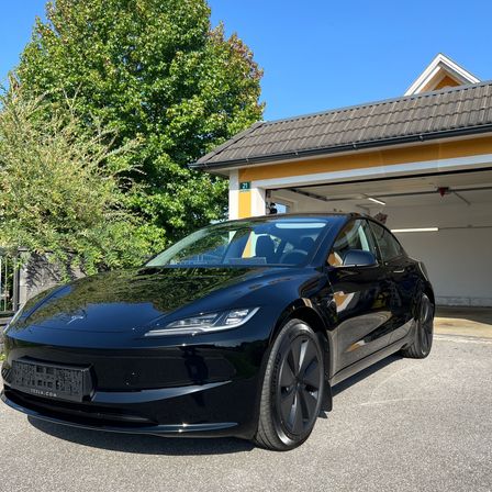 Ein schwarzer Tesla Model 3 ist vor einem Haus mit Garage geparkt. Das Auto hat ein Nummernschild und ist auf einem Betonuntergrund geparkt.