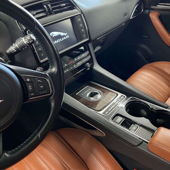 Nahaufnahme des Jaguar-Auto-Interieurs mit Ledersitzen, Lenkrad und Armaturenbrett-Steuerelementen. Das Display zeigt das Jaguar-Logo.