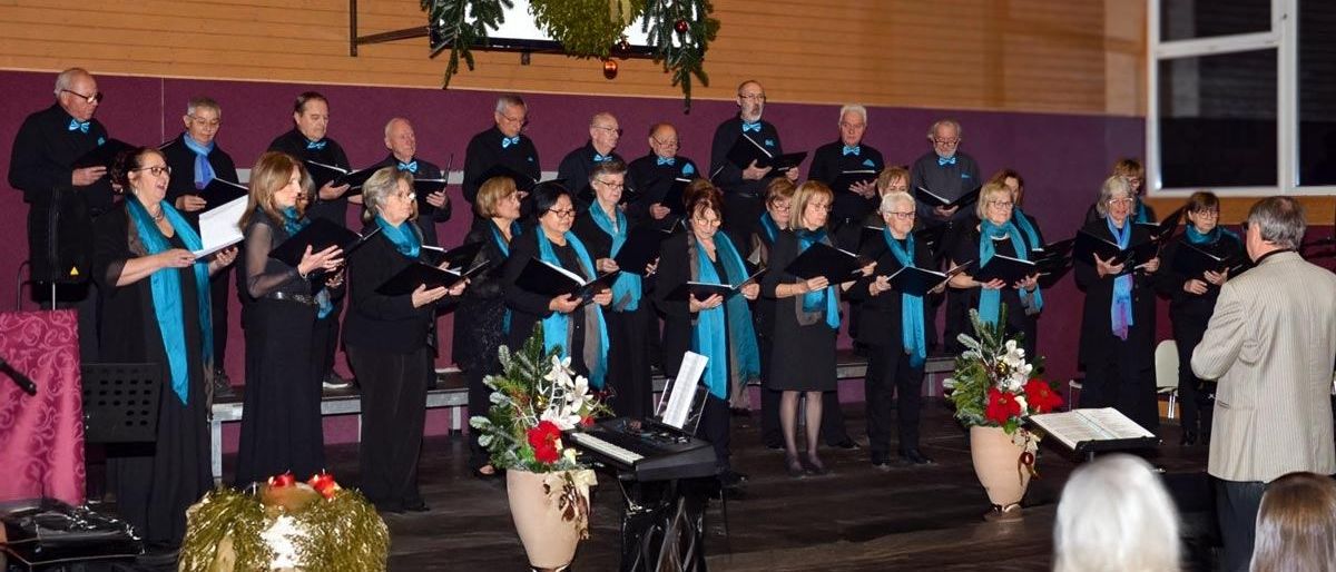 Ein Chor in Schwarz und blauen Schals tritt auf der Bühne auf und hält Liederbücher. Musikinstrumente, Blumen und Weihnachtsdekorationen zieren die Szene.