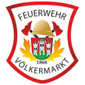 Das Emblem der Feuerwehr Volkmarkt, mit einem roten Schild, goldenem Helm und zwei Äxten, einem zentralen Wappen mit einer Burg und dem Jahr 1868 sowie einem Flammendesign.