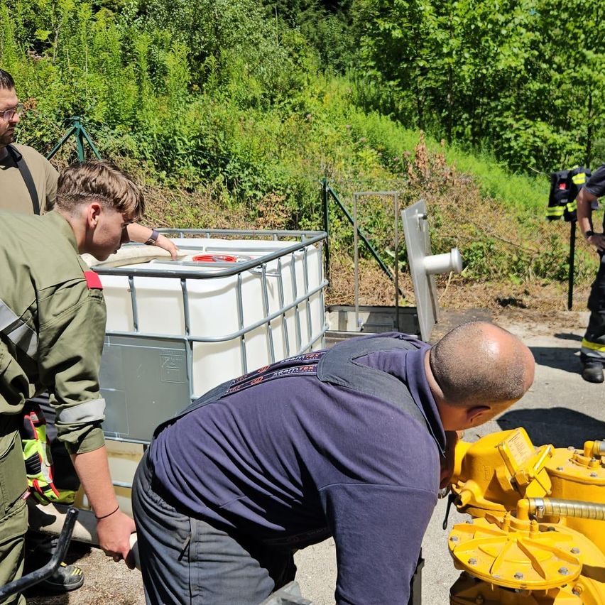 Drei Männer in Uniform arbeiten an einem gelben Ventil mit einem weißen Tank dahinter. Ein Mann schaut nach unten, während die anderen beiden zusehen.