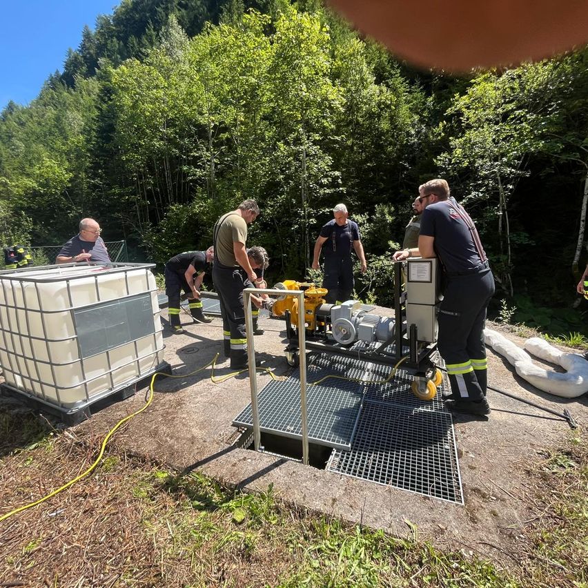Mehrere Personen in Uniformen arbeiten auf einer Betonplattform. Sie bedienen eine Pumpe und einen großen Container. Das Gebiet ist von Bäumen und Gras umgeben.