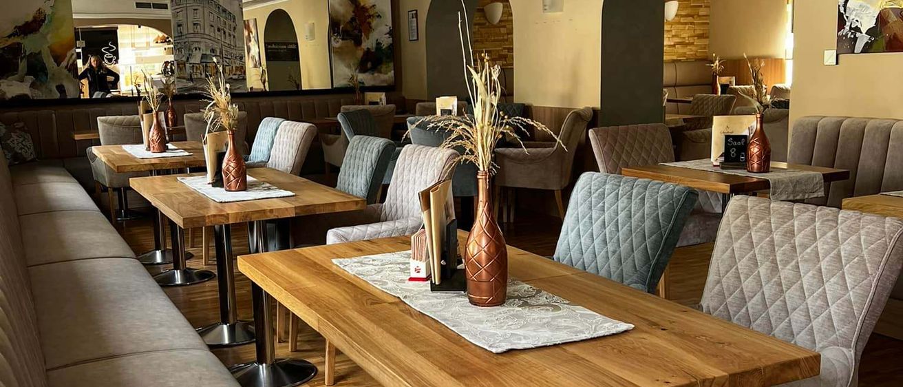 Ein modernes Restaurant mit Holztischen und blau gepolsterten Stühlen, ausgestattet mit dekorativen Vasen und Kunst an den Wänden. Der Raum hat einen großen Spiegel, der ein Gebäude reflektiert.