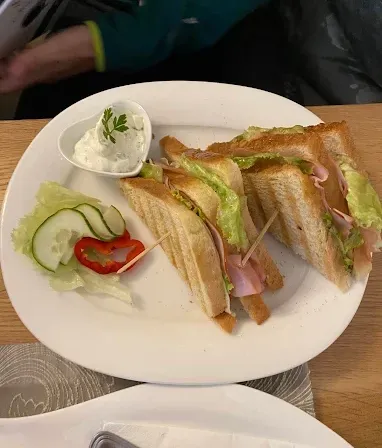 Ein weißer Teller mit zwei getoasteten Sandwiches, Gurkenscheiben, roter Paprika und einer kleinen Schüssel Dip auf einem Holztisch.