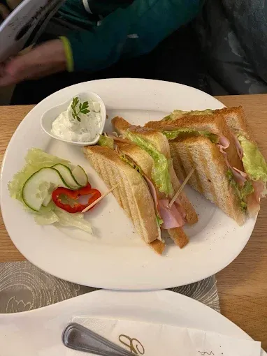 Ein weißer Teller mit zwei getoasteten Sandwiches, Gurkenscheiben, roter Paprika und einer kleinen Schüssel Dip auf einem Holztisch.