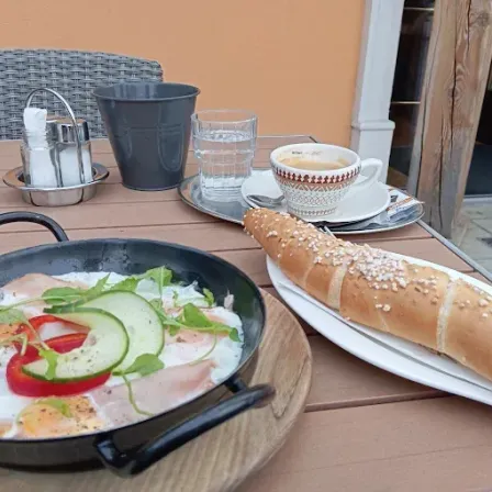 Ein Holztisch hat eine Bratpfanne mit Eiern, Gemüse und Brot. Eine Tasse Kaffee, ein Teller und ein Glas sind in der Nähe.