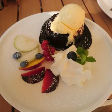 Ein weißer Teller mit einem Schokoladenkuchen, der mit Eiscreme, Schlagsahne, Beeren und geschnittenem Obst belegt ist. Der Teller ist mit Puderzucker bestreut und steht auf einem Holztisch.