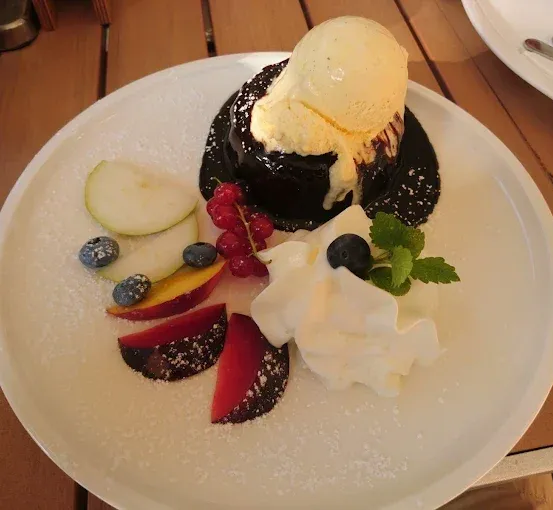 Ein weißer Teller mit einem Schokoladenkuchen, der mit Eiscreme, Schlagsahne, Beeren und geschnittenem Obst belegt ist. Der Teller ist mit Puderzucker bestreut und steht auf einem Holztisch.