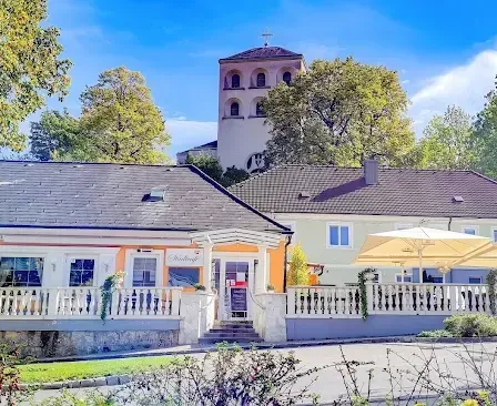 Ein malerisches Haus mit Balkon, großen Fenstern und einem Garten davor. Dahinter steht eine Kirche mit Turm und Kreuz. Bäume und Büsche umgeben das Gebiet.
