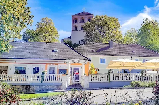 Ein malerisches Haus mit Balkon, großen Fenstern und einem Garten davor. Dahinter steht eine Kirche mit Turm und Kreuz. Bäume und Büsche umgeben das Gebiet.