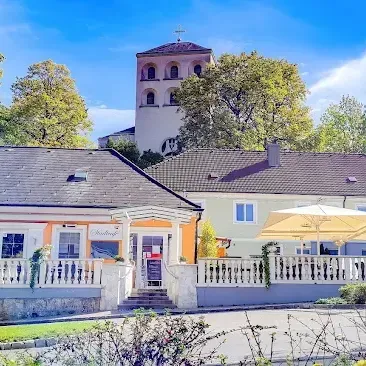 Ein zweistöckiges Gebäude mit grauem Dach und weißen Wänden steht vor einem Kirchturm. Das Gebäude hat eine Veranda mit weißen Geländern und zwei großen Sonnenschirmen. Bäume und Pflanzen umgeben das Gebäude.