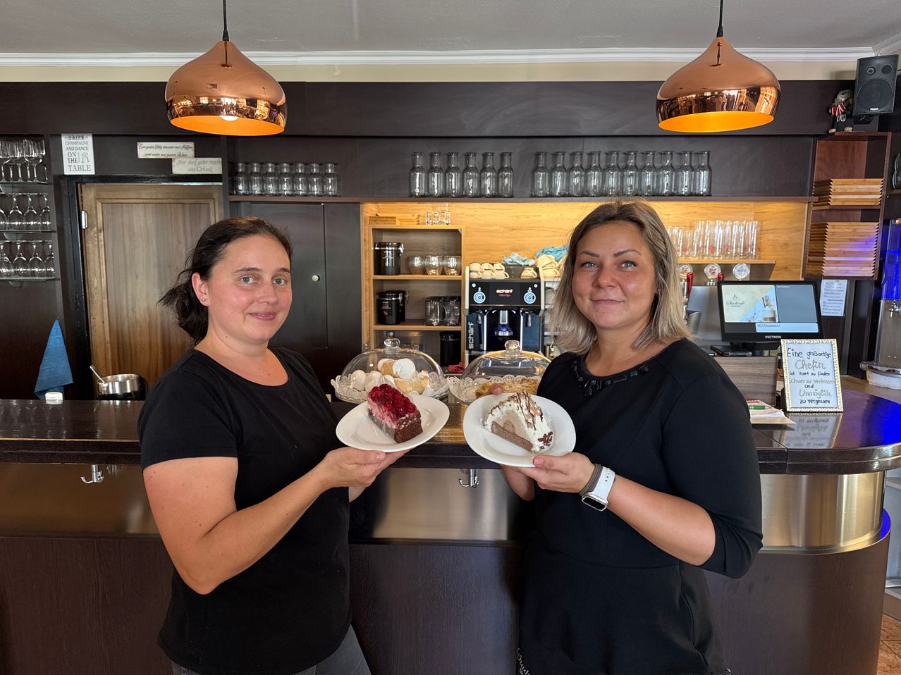 Zwei Frauen in schwarzen Shirts stehen hinter einer Theke in einem Café und halten Teller mit Desserts. Hinter ihnen steht ein hölzernes Regal mit Kaffeemaschinen, Tassen und Flaschen. Kupferne Pendelleuchten hängen von der Decke.