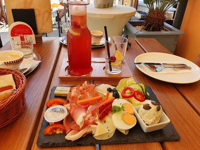 Ein Holztisch mit einem Charcuterie-Brett, einer Karaffe mit Fruchtpunsch und einem Glas. Ebenfalls auf dem Tisch stehen ein Teller mit Gabel und Messer, ein Korb mit Brot und eine Topfpflanze.