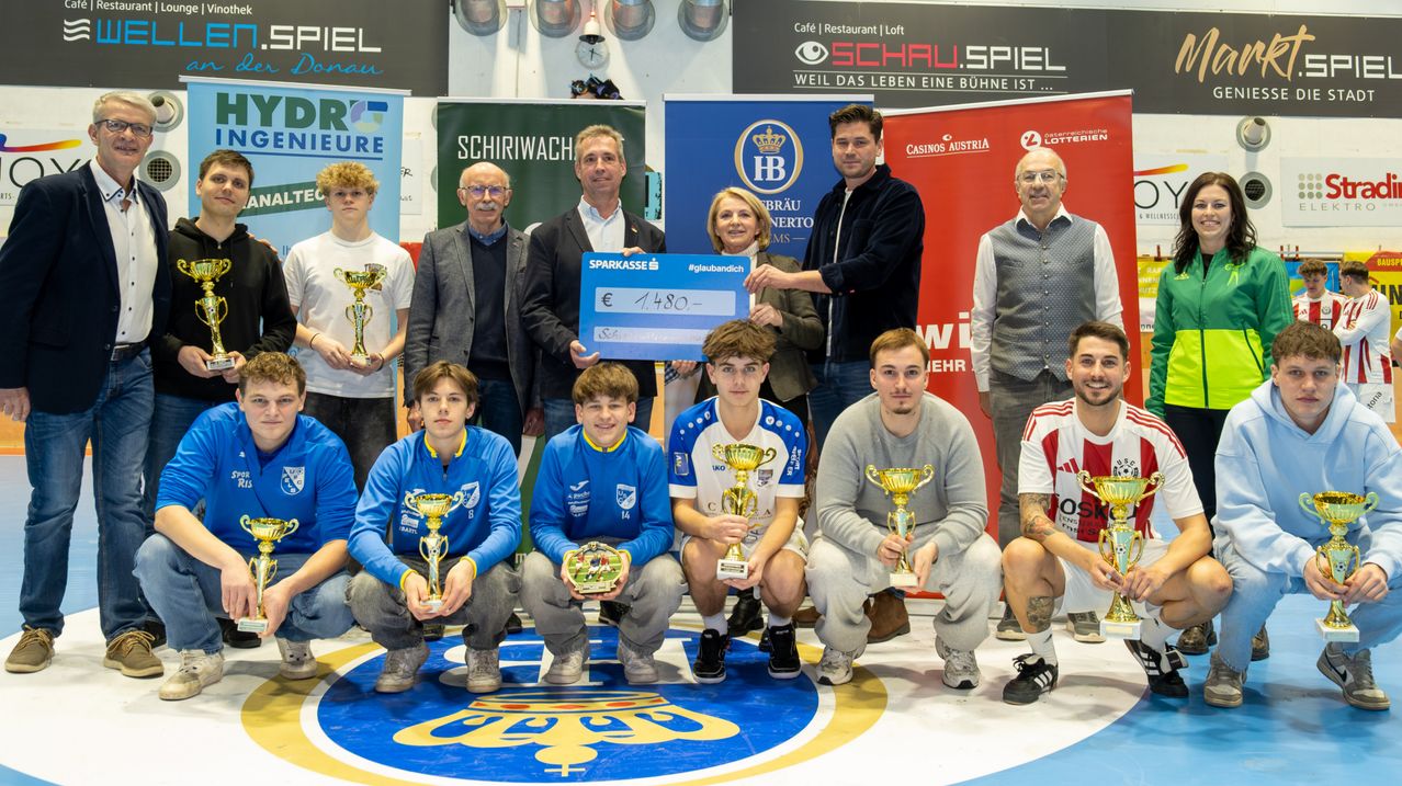 Eine Gruppe von Personen, einige mit Trophäen, posiert für ein Foto vor einem Banner. Ein Mann hält einen Scheck mit der Nummer 1480, flankiert von zwei Frauen. Dahinter stehen weitere Personen vor einem Schild.