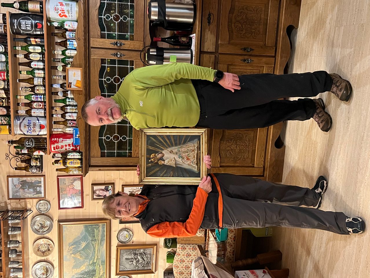 Zwei Männer stehen in einem Raum und einer hält ein gerahmtes religiöses Gemälde. Sie lächeln und posieren für ein Foto. Dahinter steht ein Schrank, der mit Flaschen und Gegenständen gefüllt ist. Mehrere gerahmte Bilder und Teller hängen an der Wand.