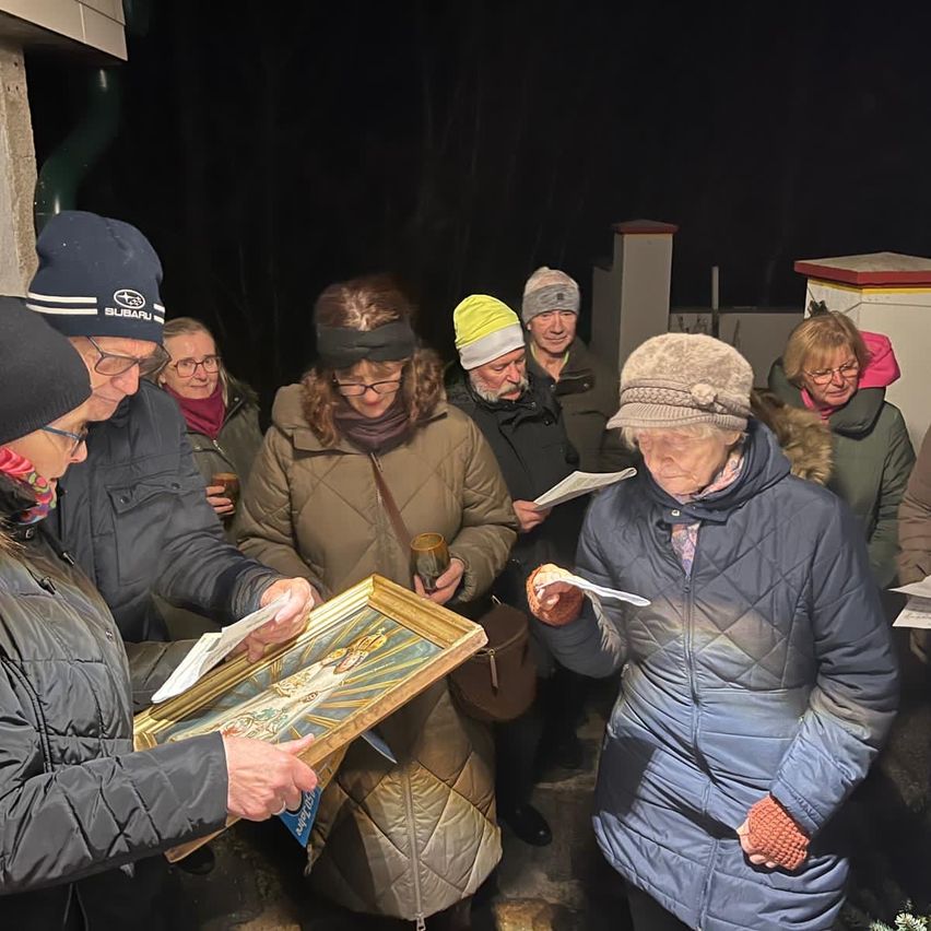 Eine Gruppe von Menschen, in Winterkleidung gekleidet, steht um ein Gebäude herum, hält ein gerahmtes Bild und liest ein Dokument. Eine Person hält ein Glas Wein.