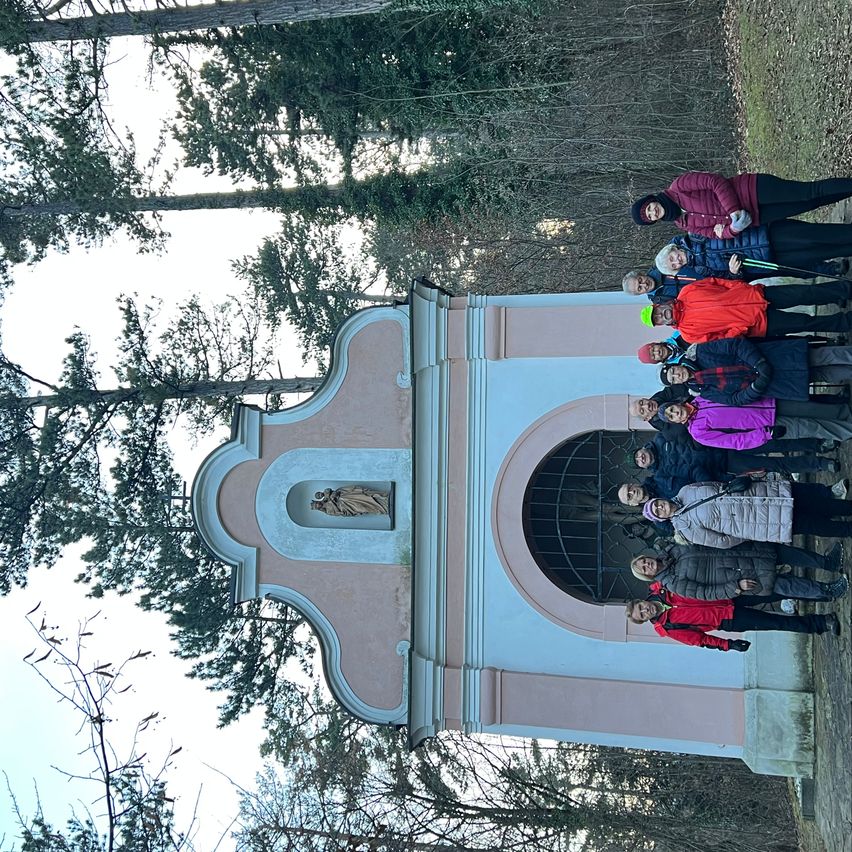 Eine Gruppe von Menschen in Winterkleidung posiert vor einer Kapelle, umgeben von Bäumen unter einem klaren Himmel.