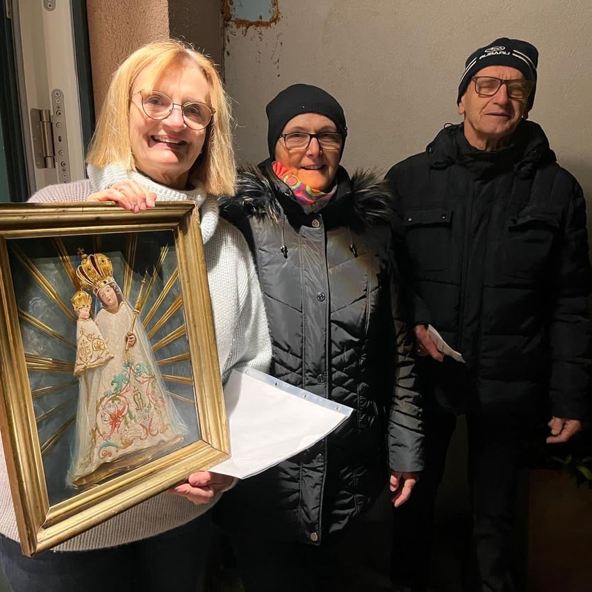 Drei Personen posieren für ein Foto mit einem gerahmten Kunstwerk, das eine gekrönte Figur darstellt. Die Frau auf der linken Seite hält den Rahmen und lächelt. Die mittlere Person trägt einen bunten Schal und eine Brille. Der Mann auf der rechten Seite hat eine schwarze Mütze und einen Mantel.