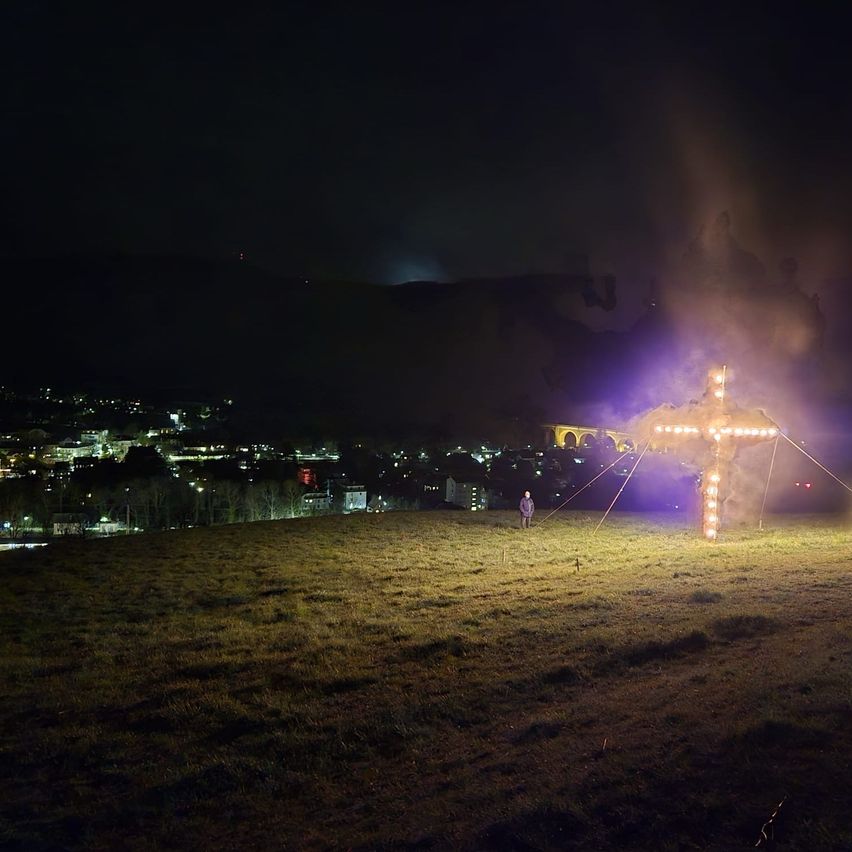 A large cross lit with lights stands in a field at night, with a city skyline in the background.