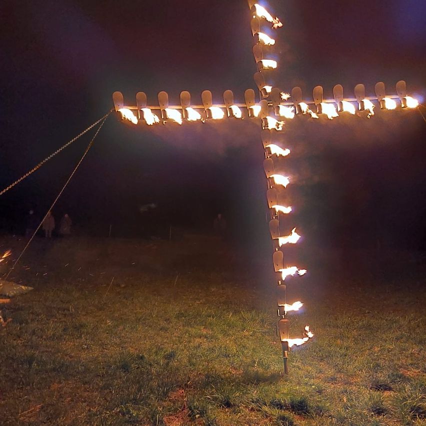 A cross made of fire with lit candles on each arm is displayed at night on a grassy field. People stand in the background.