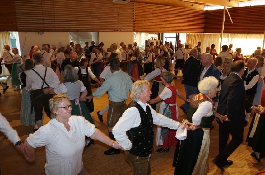 Ein Saal mit Holzboden. Eine große Gruppe von Männern und Frauen tanzt paarweise, einige in traditioneller Kleidung.