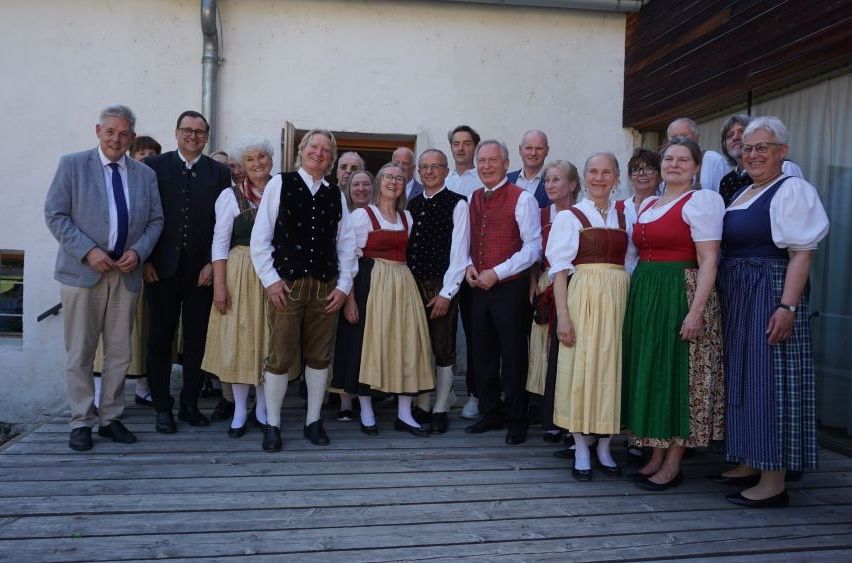 Eine Gruppe von Menschen in traditioneller Kleidung, einige in formeller Kleidung, posiert für ein Foto auf einer Holzterrasse.