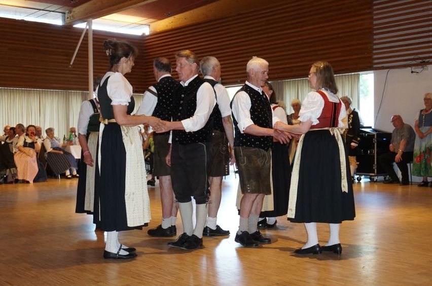 Eine Gruppe von Menschen in traditioneller Kleidung gibt sich die Hand in einer Tanzhalle. Hinter ihnen sitzen einige Leute auf Stühlen. Ein Klavier und ein Vorhang sind rechts platziert.