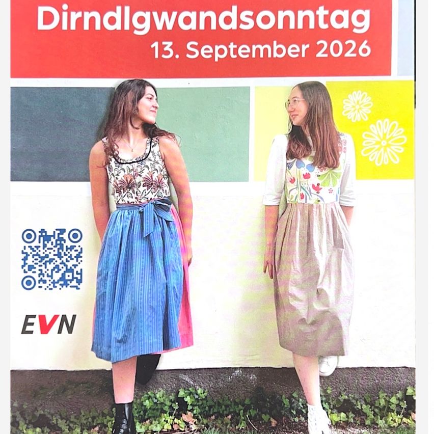 Zwei Frauen stehen vor einer bunten Wand mit einem Schild für 'Dirndlwandsonntag' am 13. September 2026. Eine trägt ein Blumenkleid und die andere ein ärmelloses Oberteil mit einem Blumenrock.