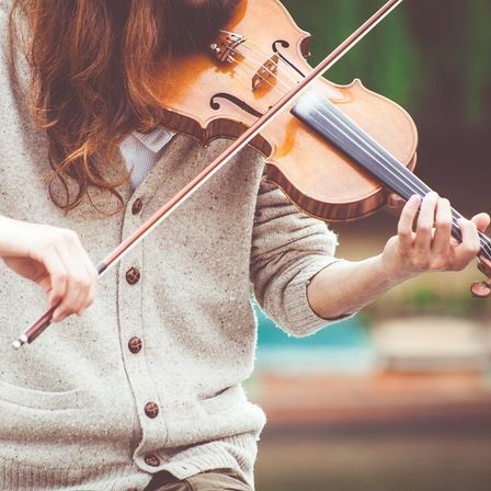 Eine Person spielt eine Geige mit einem Bogen und trägt eine graue Strickjacke. Die Geige hat einen braunen Korpus und einen schwarzen Bogen.
