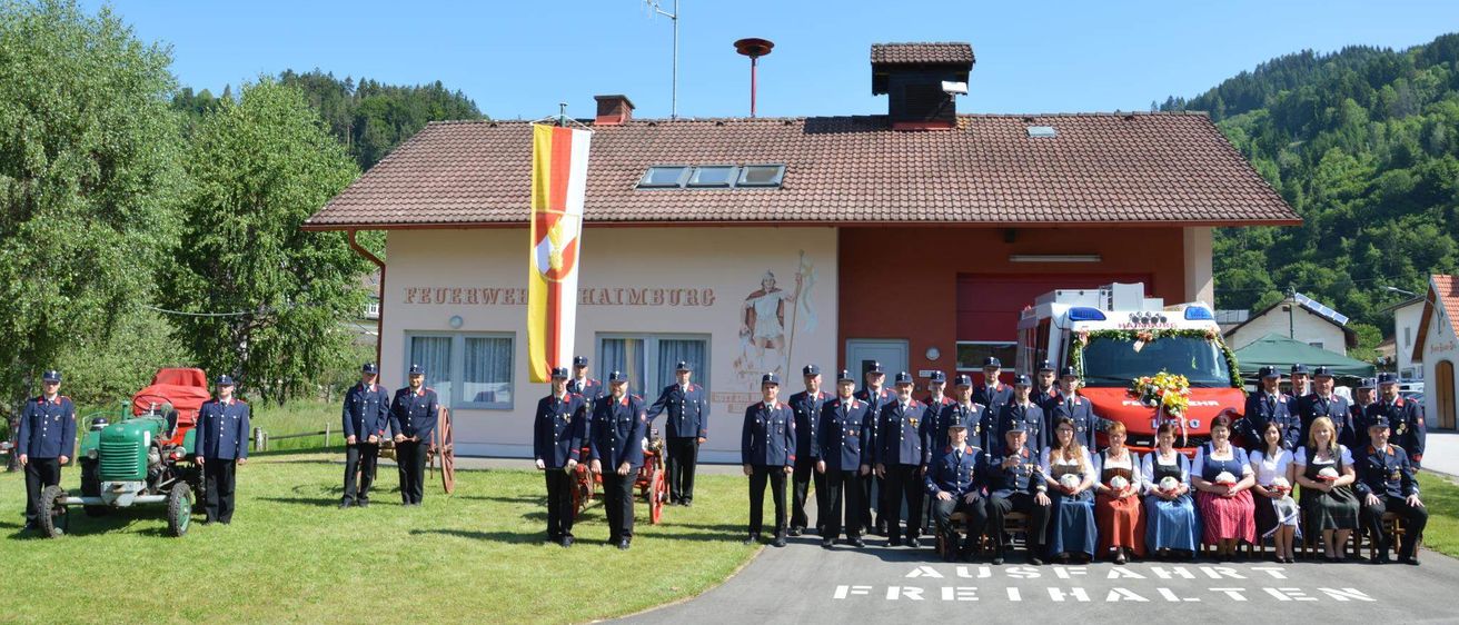 Eine Gruppe Feuerwehrleute in Uniform posiert für ein Foto vor einer Feuerwache mit einer Flagge und einem Feuerwehrauto im Hintergrund.