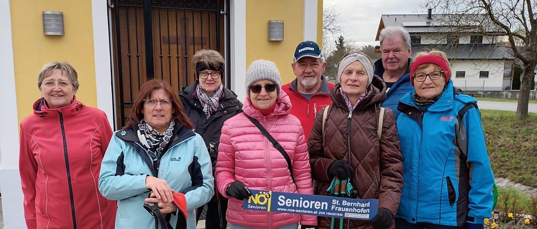 Sieben Senioren posieren für ein Foto vor einem Gebäude. Einer von ihnen hält ein Schild mit der Aufschrift 'Senioren'. Die Frau links hält die Leine eines Hundes.