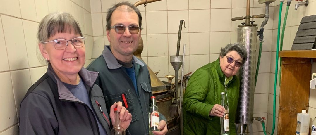 Drei Personen stehen in einem Raum, zwei halten Flaschen und eine gießt Flüssigkeit in einen Trichter.