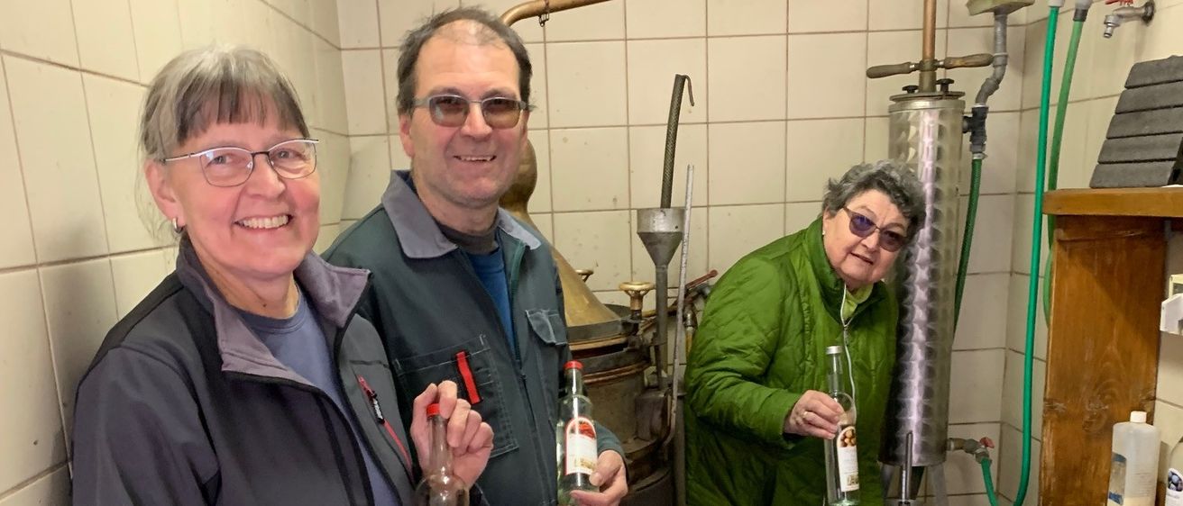 Drei Personen stehen in einem Raum, zwei halten Flaschen und eine gießt Flüssigkeit in einen Trichter.