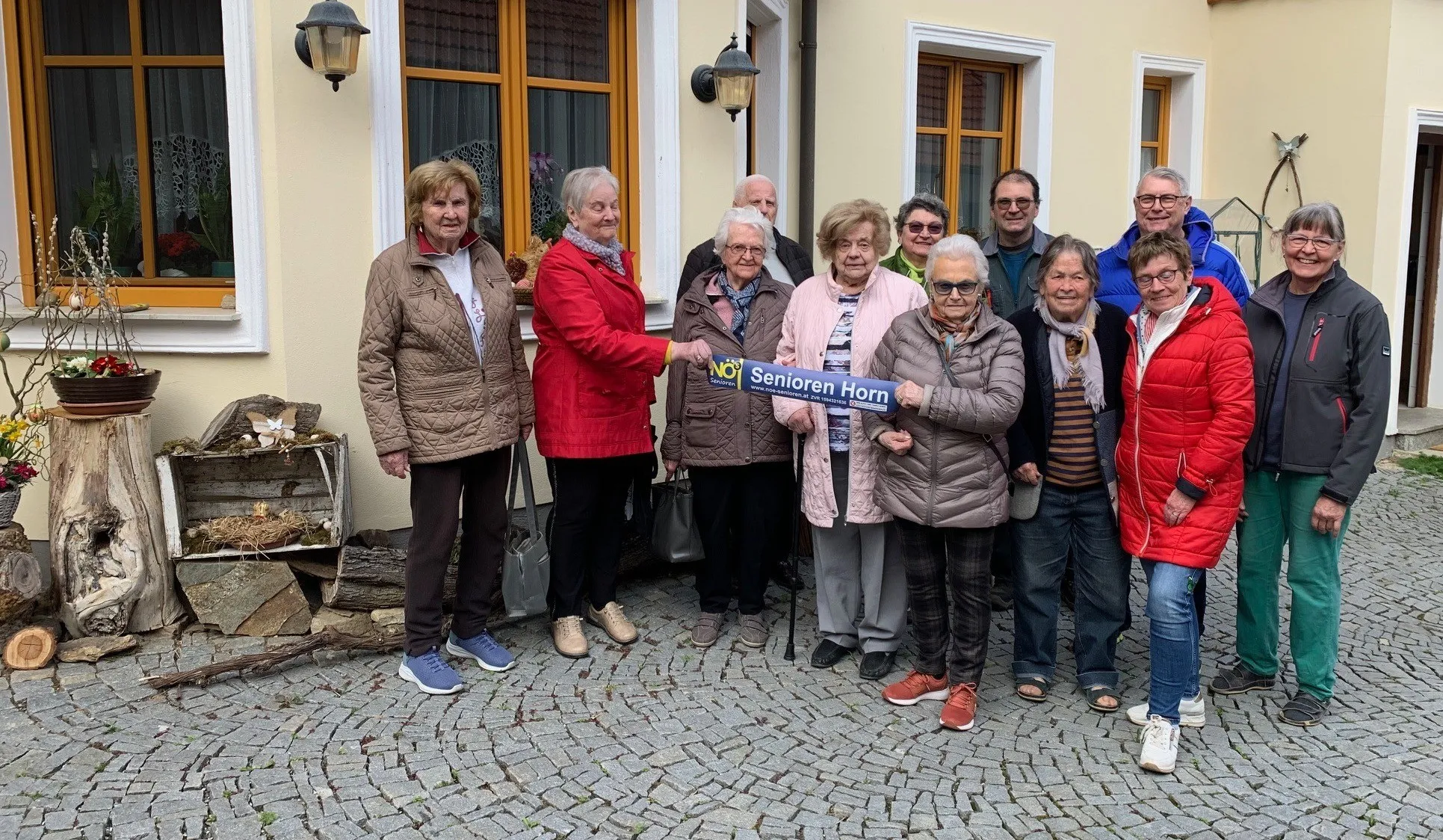 Eine Gruppe älterer Personen, die lächelnd für ein Foto posieren, mit einer Frau, die ein blaues Banner mit der Aufschrift 'Senioren Horn' hält. Sie stehen vor einem Gebäude mit Fenstern und einer Lampe an der Wand.