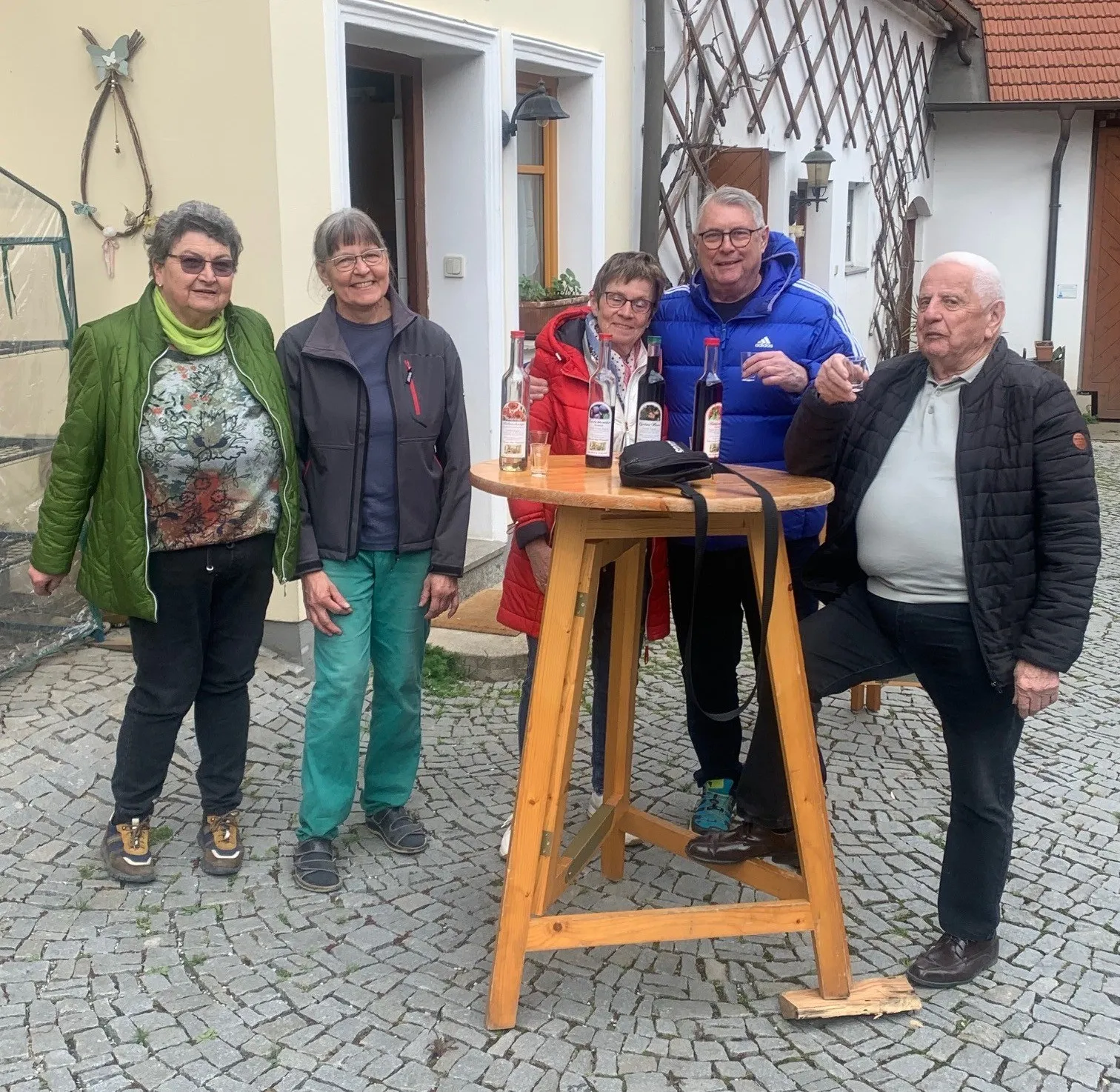 Fünf ältere Erwachsene stehen um einen kleinen Holztisch mit Flaschen und Gläsern. Sie sind vor einem Gebäude mit einer Lampe.
