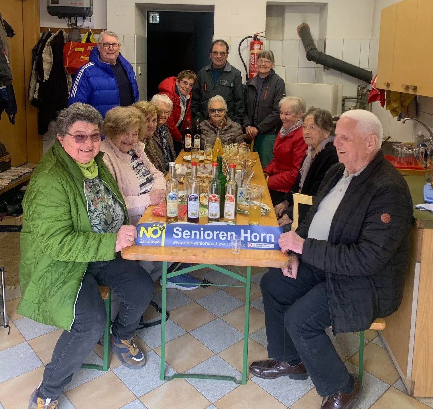 Eine Gruppe älterer Menschen sitzt um einen Tisch herum, lächelt und posiert für ein Foto. Der Tisch ist mit Flaschen und Gläsern geschmückt, und eine Banner mit der Aufschrift 'Senioren Horn' ist darauf angebracht.