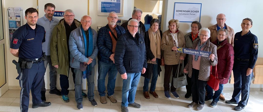 Eine Gruppe von Menschen steht in einem Raum mit gefliesten Böden und weißen Wänden. Sie posieren für ein Foto. Einige von ihnen halten ein blaues Schild. Es gibt Plakate an der Wand.