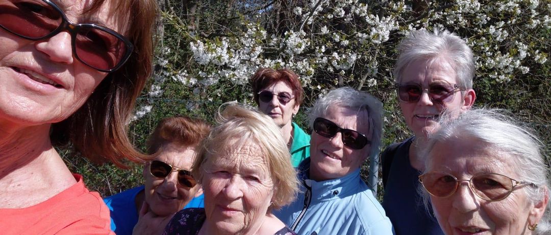 Eine Gruppe älterer Frauen steht im Freien, lächelt und posiert für ein Foto vor einem blühenden Baum.