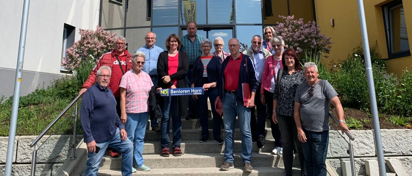 Eine Gruppe von Erwachsenen steht auf Stufen vor einem Gebäude mit gelben Fahnen, einem blauen Schild und einer Glaswand. Der Himmel ist klar mit einigen Wolken.