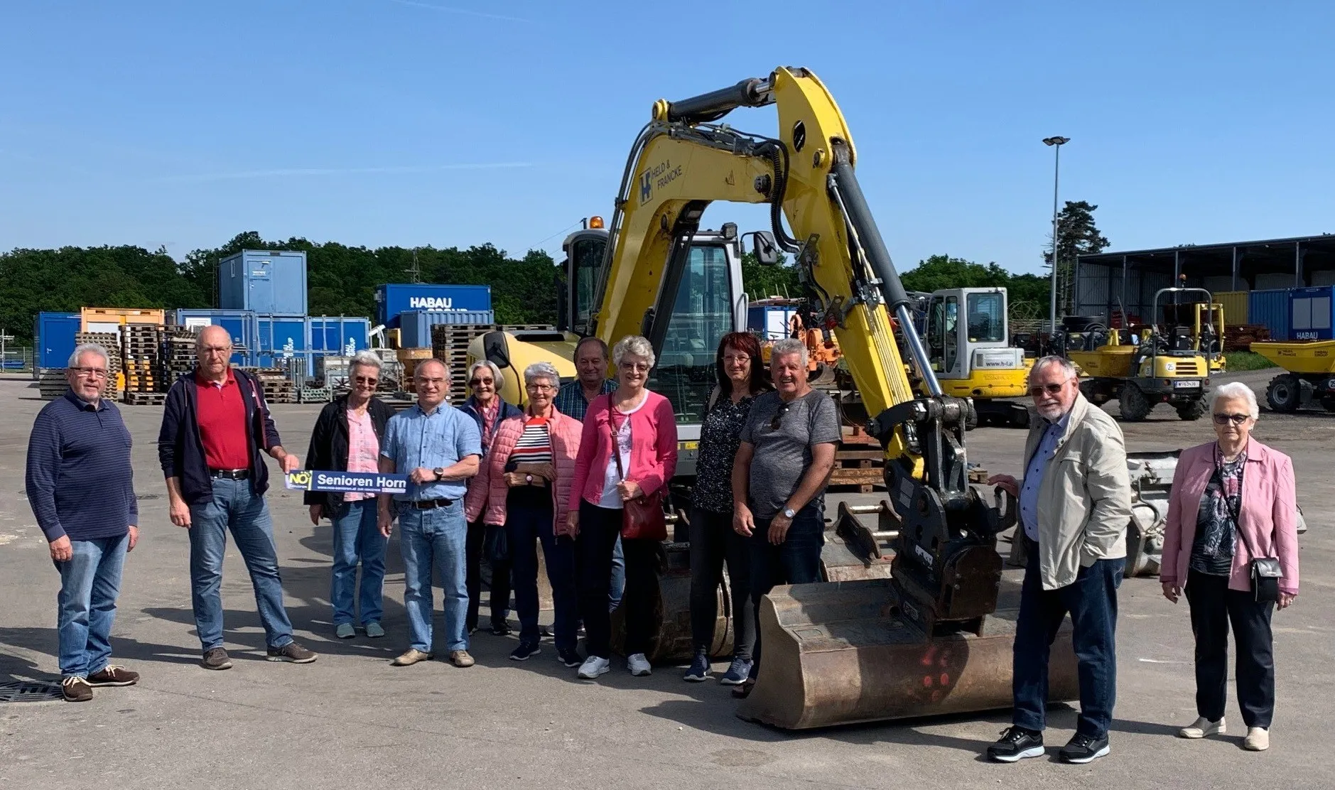 Eine Gruppe von Menschen steht vor einem gelben Bagger und posiert für ein Foto. Sie tragen alle Freizeitkleidung. Hinter ihnen ist mehr Maschinen geparkt, und in der Ferne sind Bäume und ein klarer blauer Himmel zu sehen.