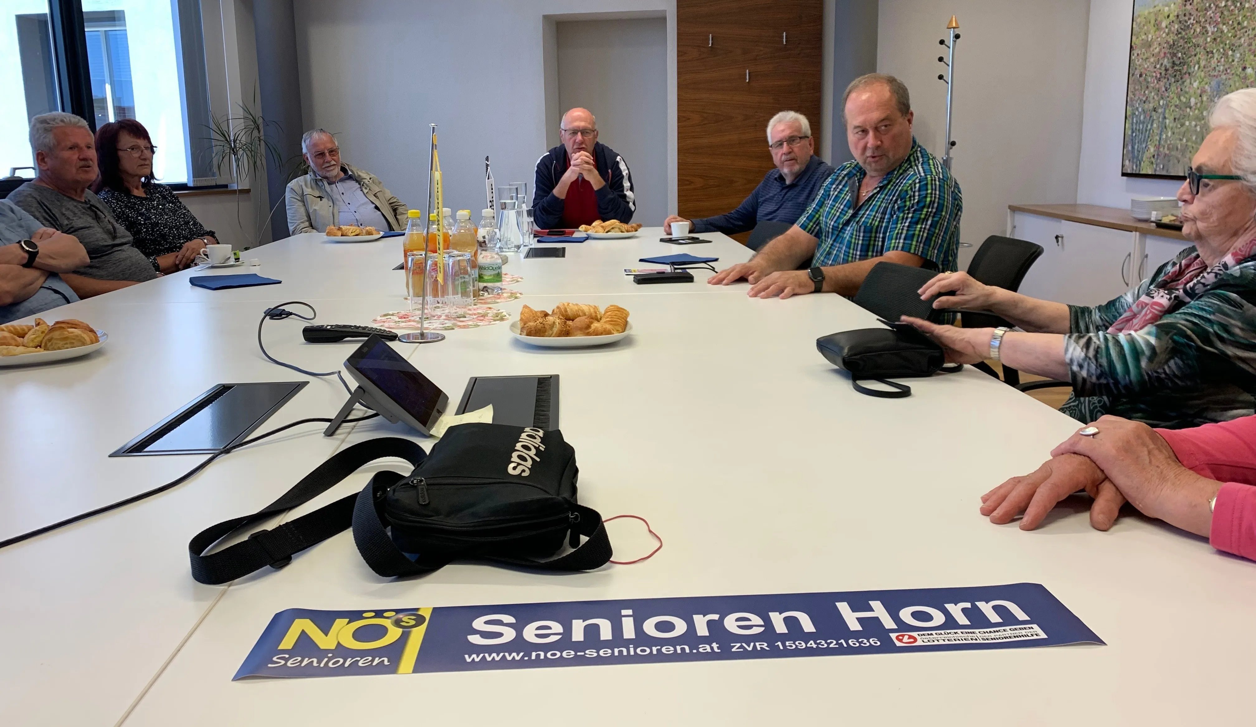 Fünf Männer in einem Besprechungsraum mit einem Tisch voller Essen und Getränken. Ein Banner mit dem Text 'Senioren Horr' ist unten.