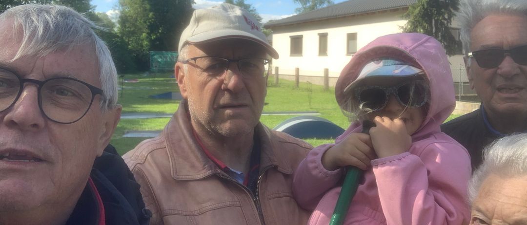 Drei Menschen stehen draußen in der Nähe eines Hauses. Ein älterer Mann trägt eine Baseball-Kappe und eine Brille. Ein junges Mädchen hält einen grünen Schlauch und trägt eine rosa Regenjacke. Ein weiterer Mann steht neben ihnen. Bäume und ein Feld sind hinter ihnen.
