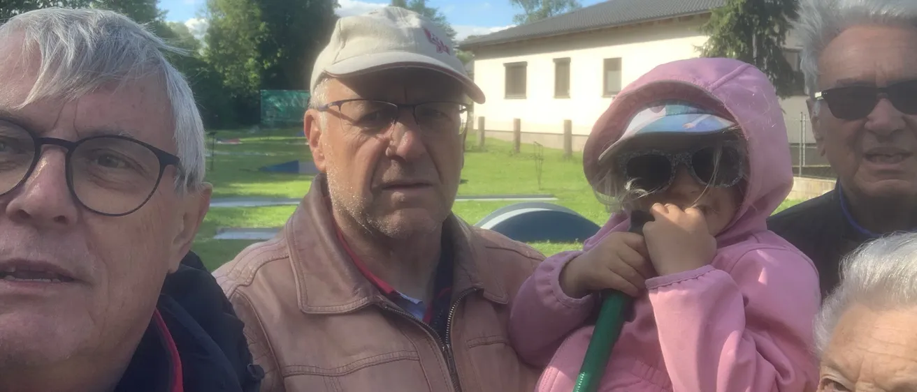 Drei Menschen stehen draußen in der Nähe eines Hauses. Ein älterer Mann trägt eine Baseball-Kappe und eine Brille. Ein junges Mädchen hält einen grünen Schlauch und trägt eine rosa Regenjacke. Ein weiterer Mann steht neben ihnen. Bäume und ein Feld sind hinter ihnen.