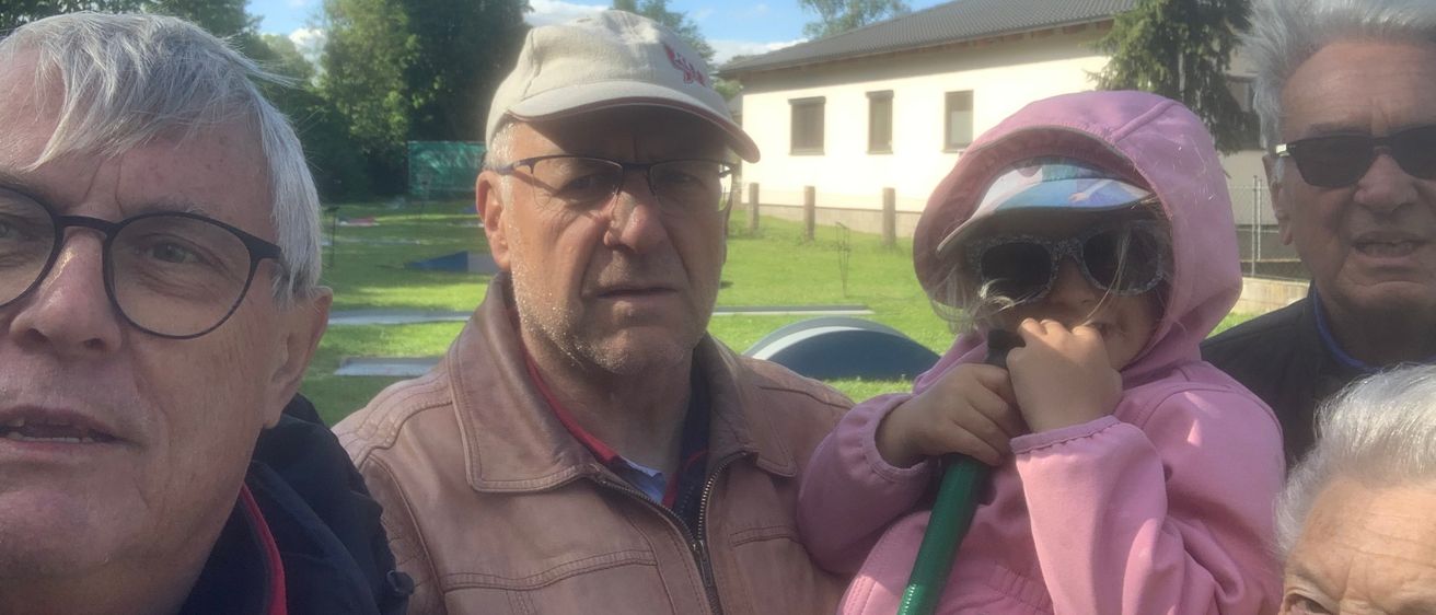 Drei Menschen stehen draußen in der Nähe eines Hauses. Ein älterer Mann trägt eine Baseball-Kappe und eine Brille. Ein junges Mädchen hält einen grünen Schlauch und trägt eine rosa Regenjacke. Ein weiterer Mann steht neben ihnen. Bäume und ein Feld sind hinter ihnen.