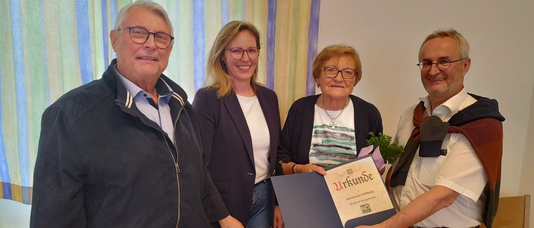 Eine Gruppe von vier Erwachsenen steht vor einer weißen Wand mit einem Vorhang. Die Frau in der Mitte hält ein Zertifikat, und der Mann rechts trägt einen Anzug. Die Frau links trägt eine Brille und eine Halskette.