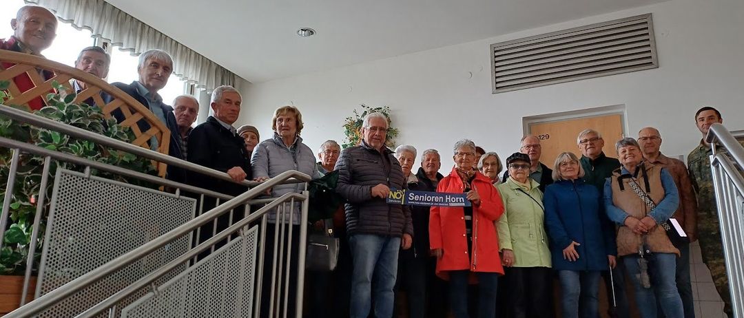 Eine Gruppe älterer Personen posiert für ein Foto in einem Gebäude mit einem blauen Schild, das Senioren Horn sagt.