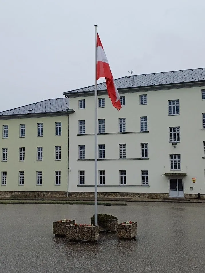 Eine rot-weiße Flagge weht auf einem Mast vor einem Gebäude mit vielen Fenstern und einem grauen Dach.
