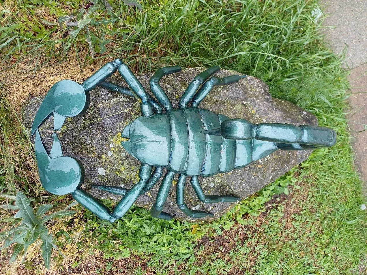 Eine grüne Skorpionsskulptur sitzt auf einem Felsen in einer Grasfläche. Die Skulptur ist detailliert gestaltet und hat einen metallischen Glanz.