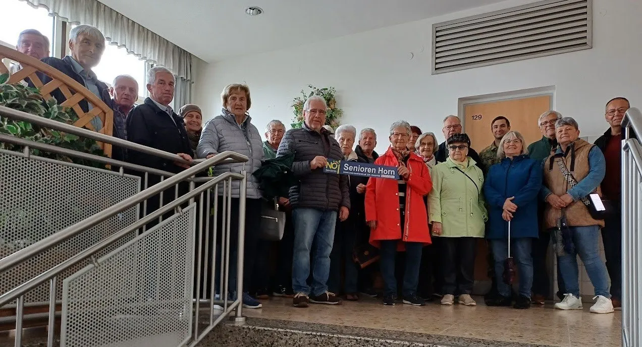 Eine Gruppe älterer Menschen steht zusammen und hält ein Schild mit der Aufschrift 'Senioren Horn'. Sie tragen alle Winterkleidung. Hinter ihnen ist eine Wand mit einer Tür und einem Fenster.