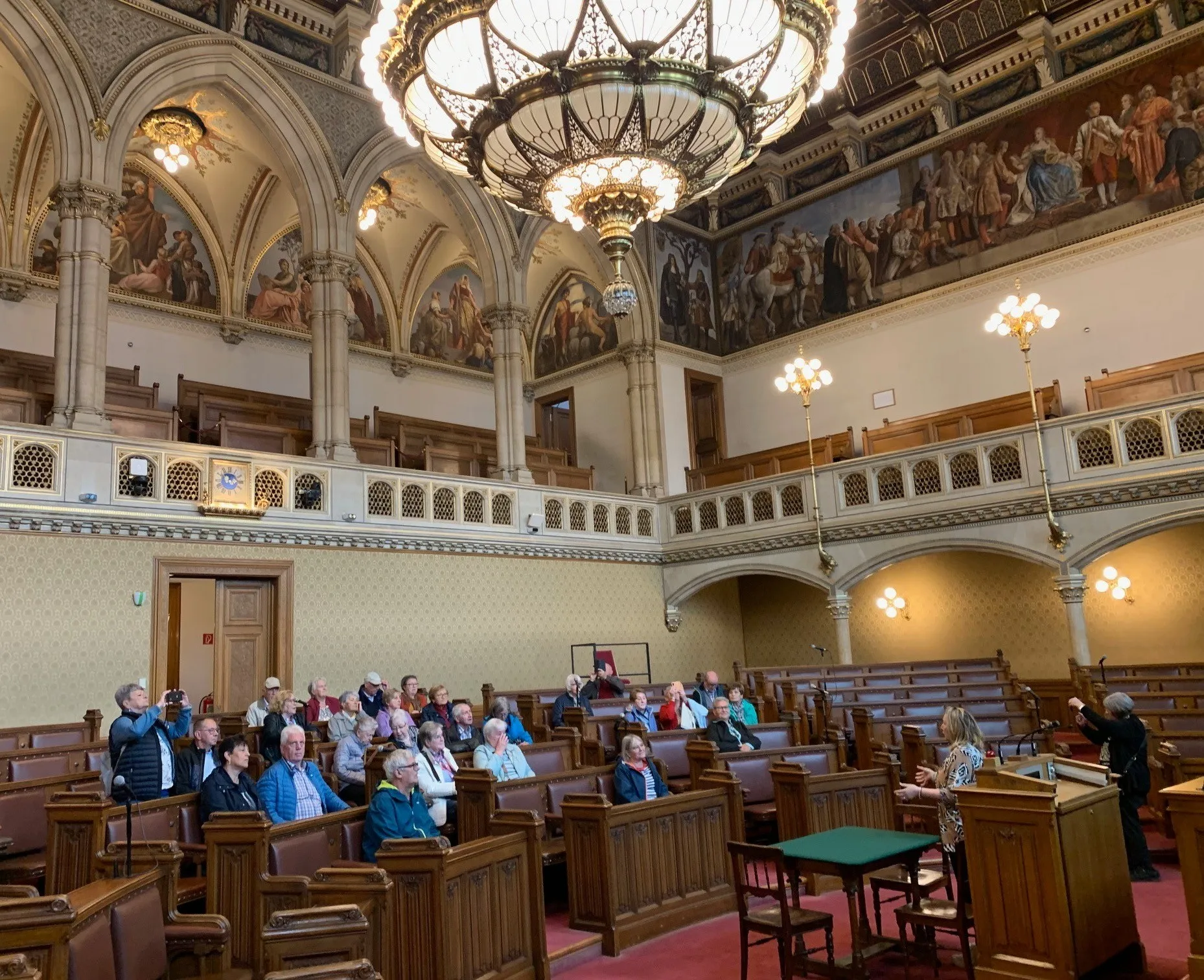 Ein großer Saal mit gewölbten Decken und hölzernen Kirchenbänken. Eine große Kronleuchter hängt von der Decke. Eine Frau steht an einem Rednerpult und spricht zu einer Gruppe sitzender Personen. Historische Gemälde schmücken die Wände.
